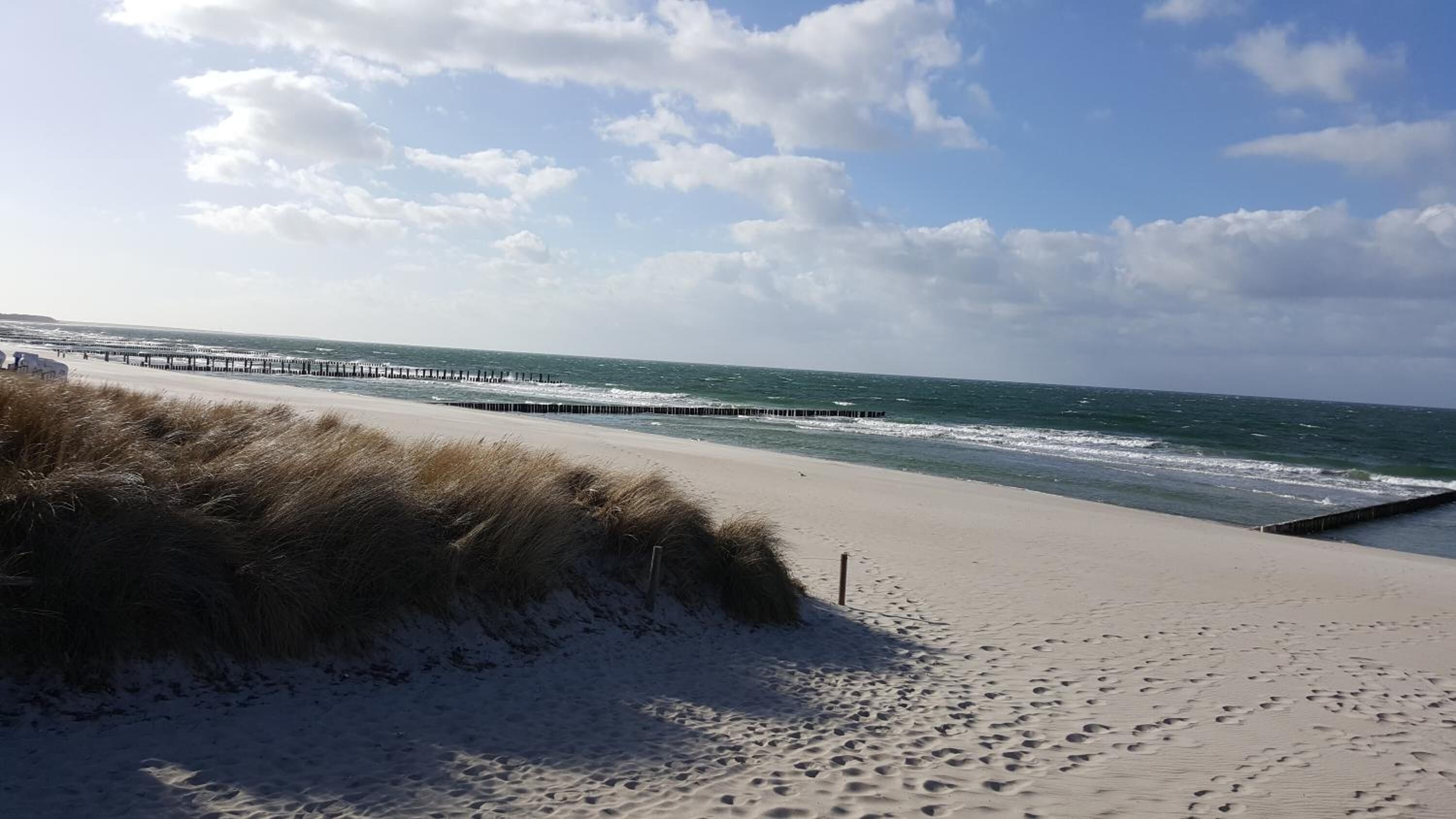 Ein Strand mit einem hölzernen Steg, der sich ins Meer erstreckt, Sanddünen und Fußabdrücken im Sand.