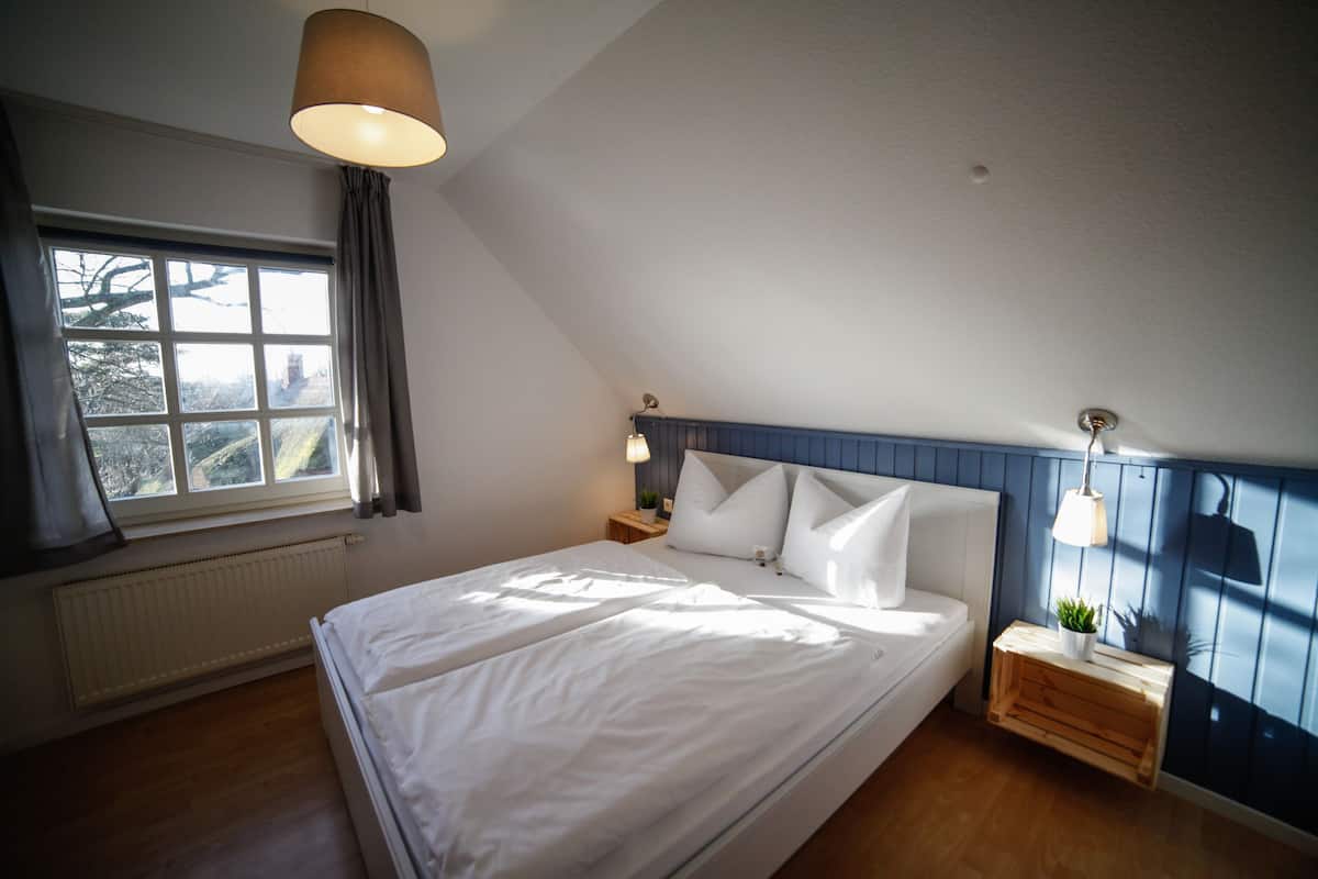 A bedroom with a large bed, a window with curtains, a bedside table with a lamp, and a wall-mounted plant.