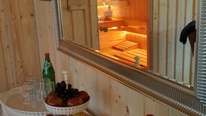 Interior em madeira com um espelho refletindo um espaço para sauna. Há um prato de frutas, uma garrafa e copos sobre uma mesa.