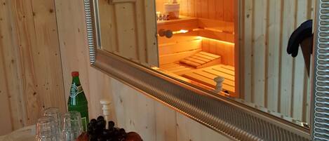 A wooden interior with a mirror reflecting a sauna room. There is a fruit platter, a bottle, and glasses on a table.