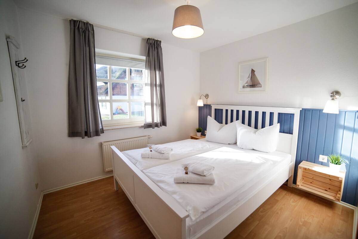 A double bed with white linens, a bedside table with a lamp, a window with curtains, and a small plant on a wooden side table.