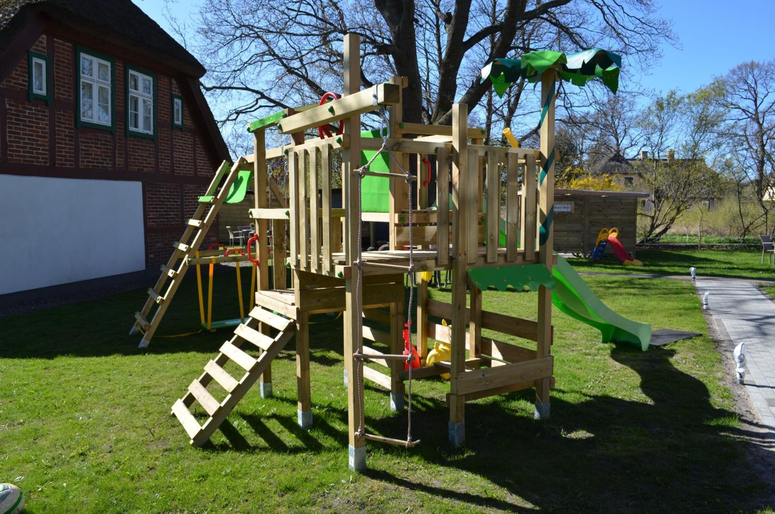 Una estructura de juego de madera con toboganes y elementos para escalar, ubicada en un área verde con un edificio de ladrillo al fondo.