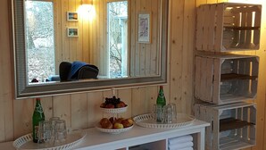 Interior de uma cabana de madeira com um espelho grande, um console branco com toalhas e revistas, e uma prateleira na parede com copos e garrafas.