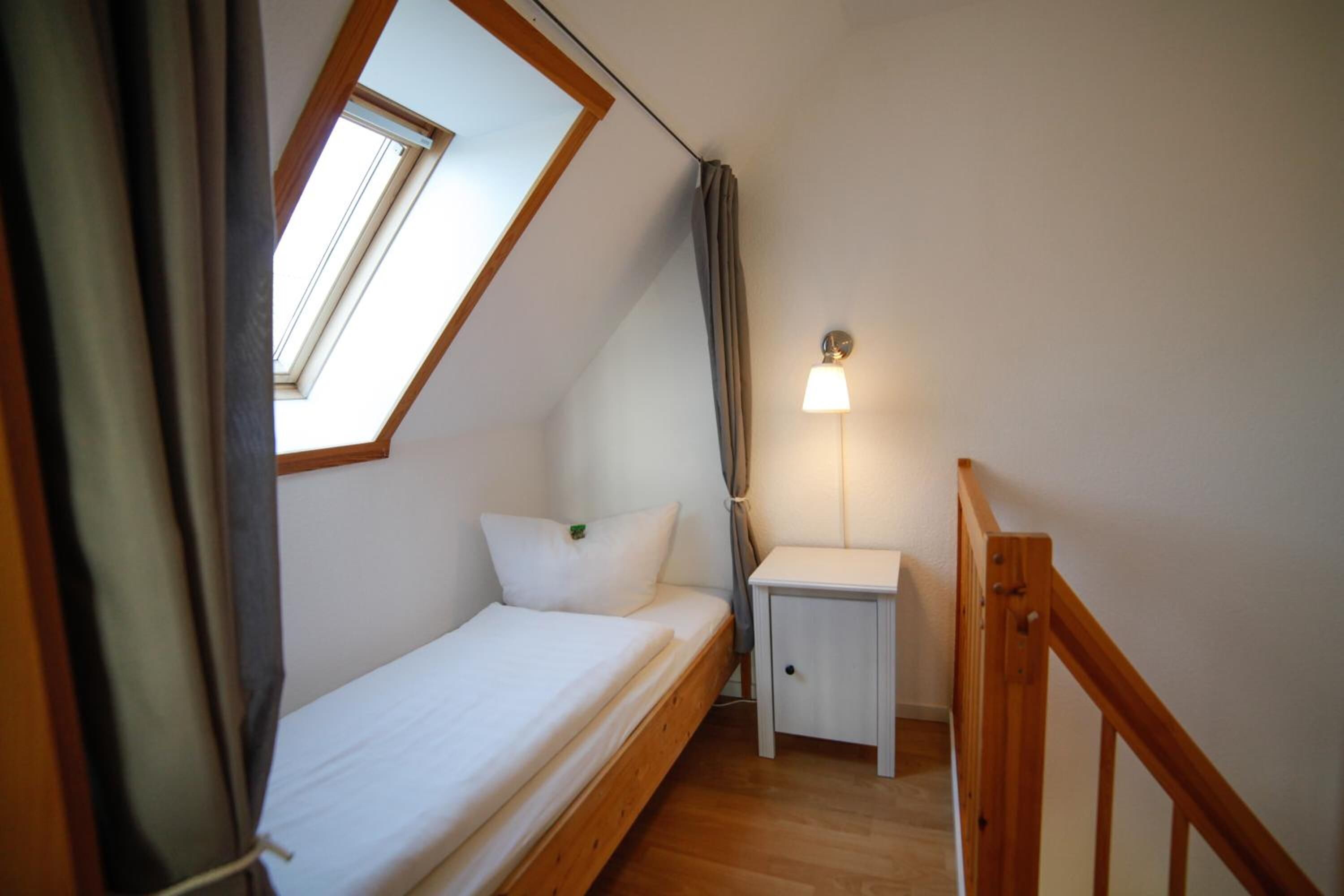 Une petite pièce bien éclairée, avec un lit simple, une table de chevet, une commode et une fenêtre avec des rideaux.