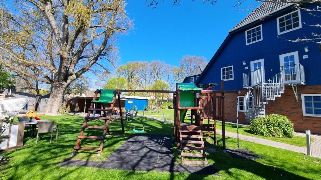 A playground with a swing set and climbing structures, a brick house with a blue exterior, and a large tree in the background.