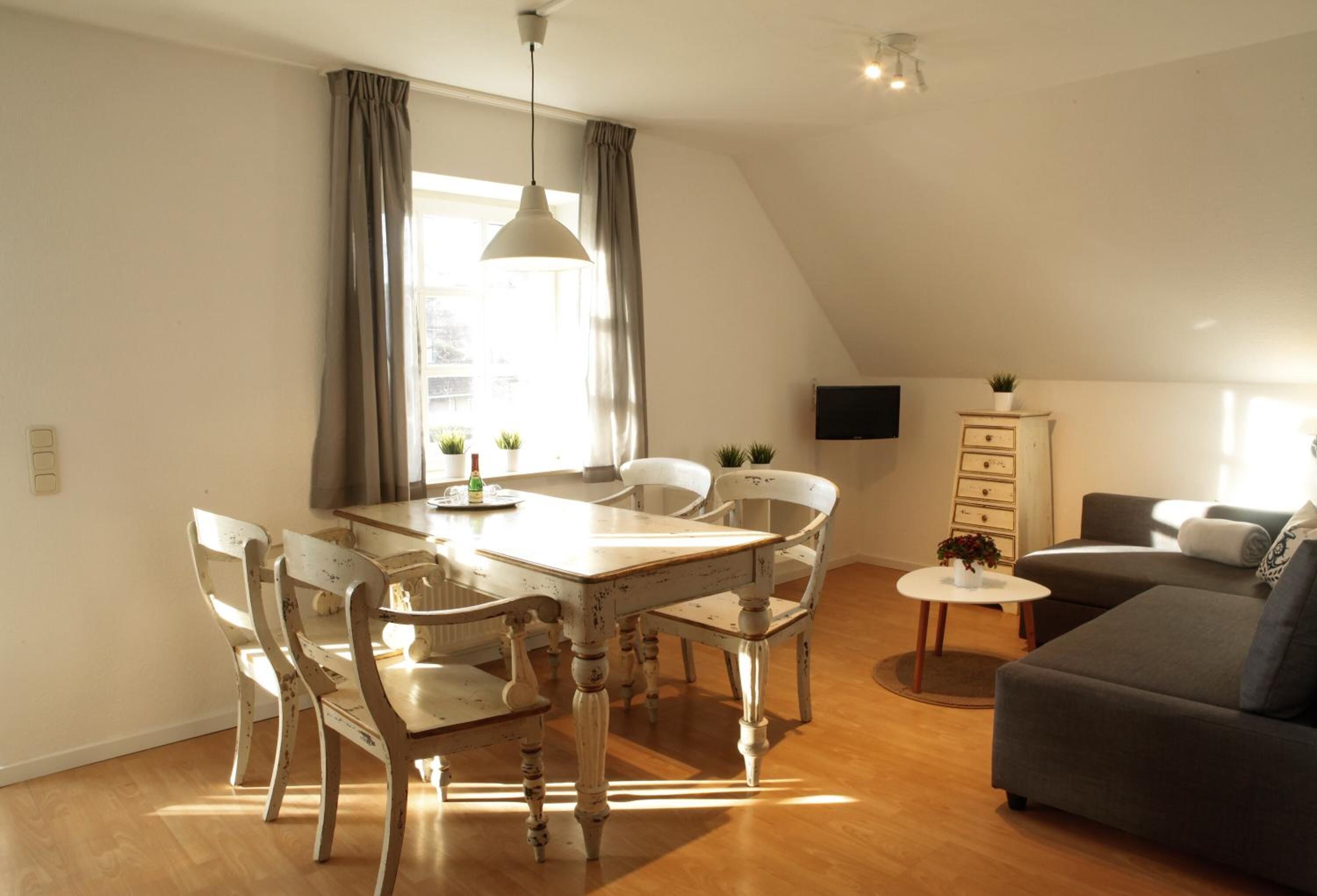 Un comedor con mesa y sillas de madera, una mesita redonda con una planta, un sofá gris y un televisor en la pared.