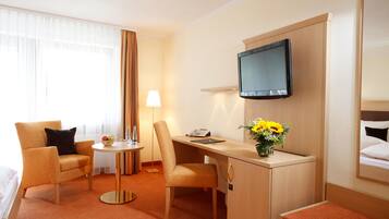 Ein Hotelzimmer mit Schreibtisch, zwei Stühlen, Fernseher, Lampe, einem kleinen Tisch mit Blumenvase und einem Bett mit Kopfteil.