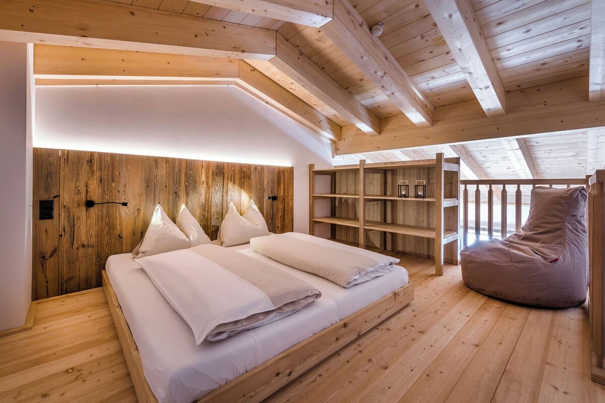 A bedroom with a wooden ceiling, a bed with white bedding, a bean bag chair, and built-in wooden shelves.