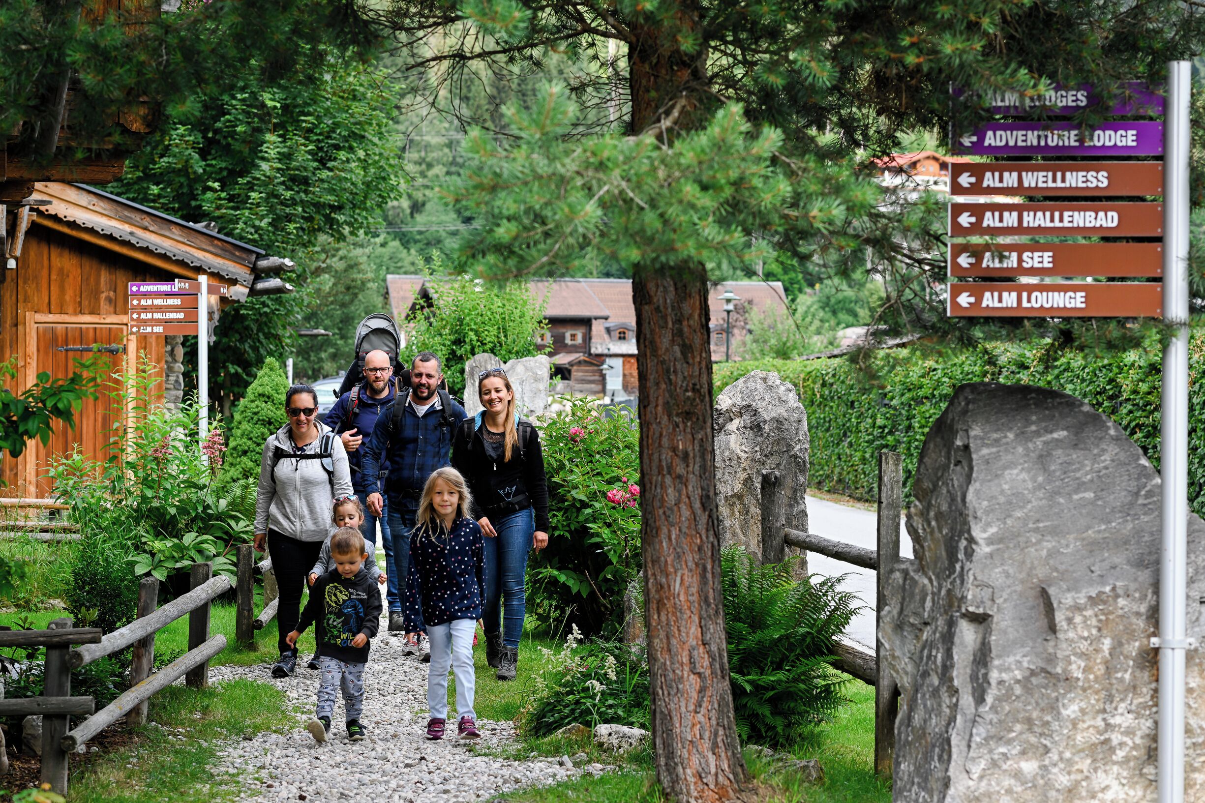 Eine Familie geht auf eine Holzhütte zu, an deren Schild Wegweiser zu verschiedenen Einrichtungen wie Adventure Lodge, Alm Wellness, Alm Hallenbad, Alm See und Alm Lounge zu sehen sind.