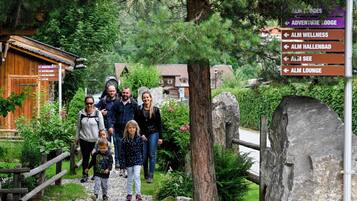 Eine Familie geht auf eine Holzhütte zu, an deren Schild Wegweiser zu verschiedenen Einrichtungen wie Adventure Lodge, Alm Wellness, Alm Hallenbad, Alm See und Alm Lounge zu sehen sind.