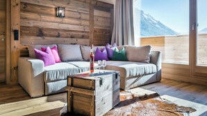 A cozy room with a wooden wall, a sofa with colorful cushions, a rustic chest of drawers, and a view of a snowy mountain. - Almdorf Almlust (Flachau)