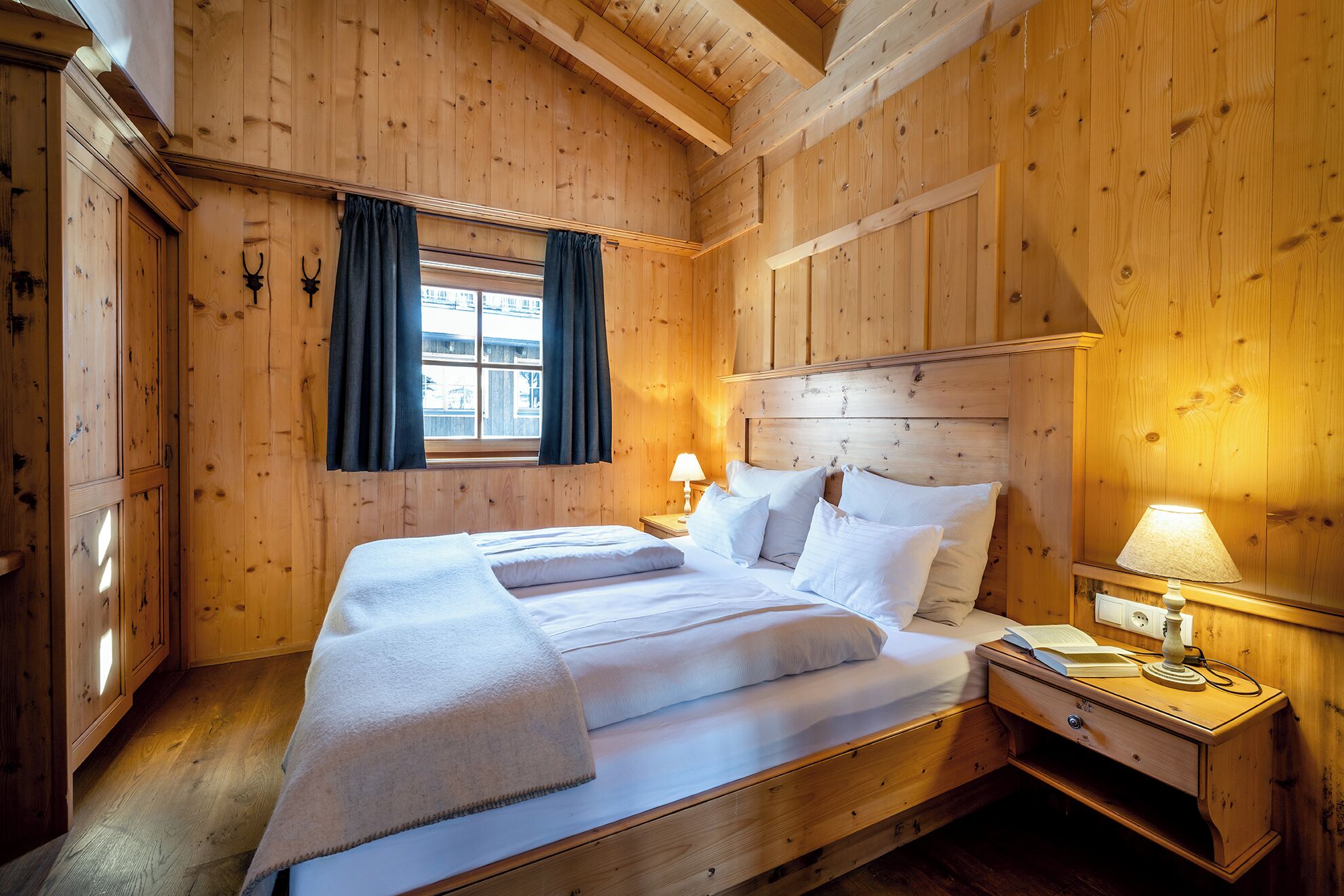 A wooden room with a bed, a nightstand, a lamp, and a window with curtains.