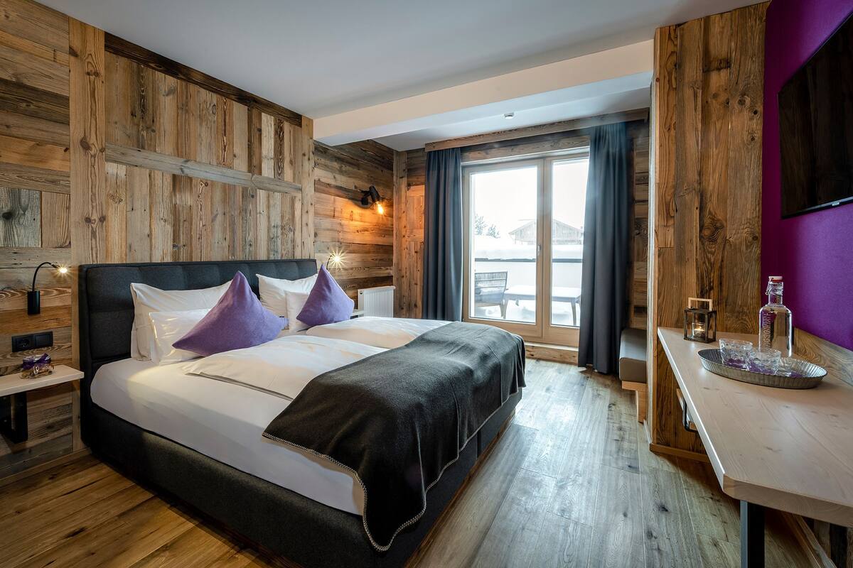 A hotel room with a wooden wall, a bed with white bedding, a nightstand, a desk with a tray, and a large window.