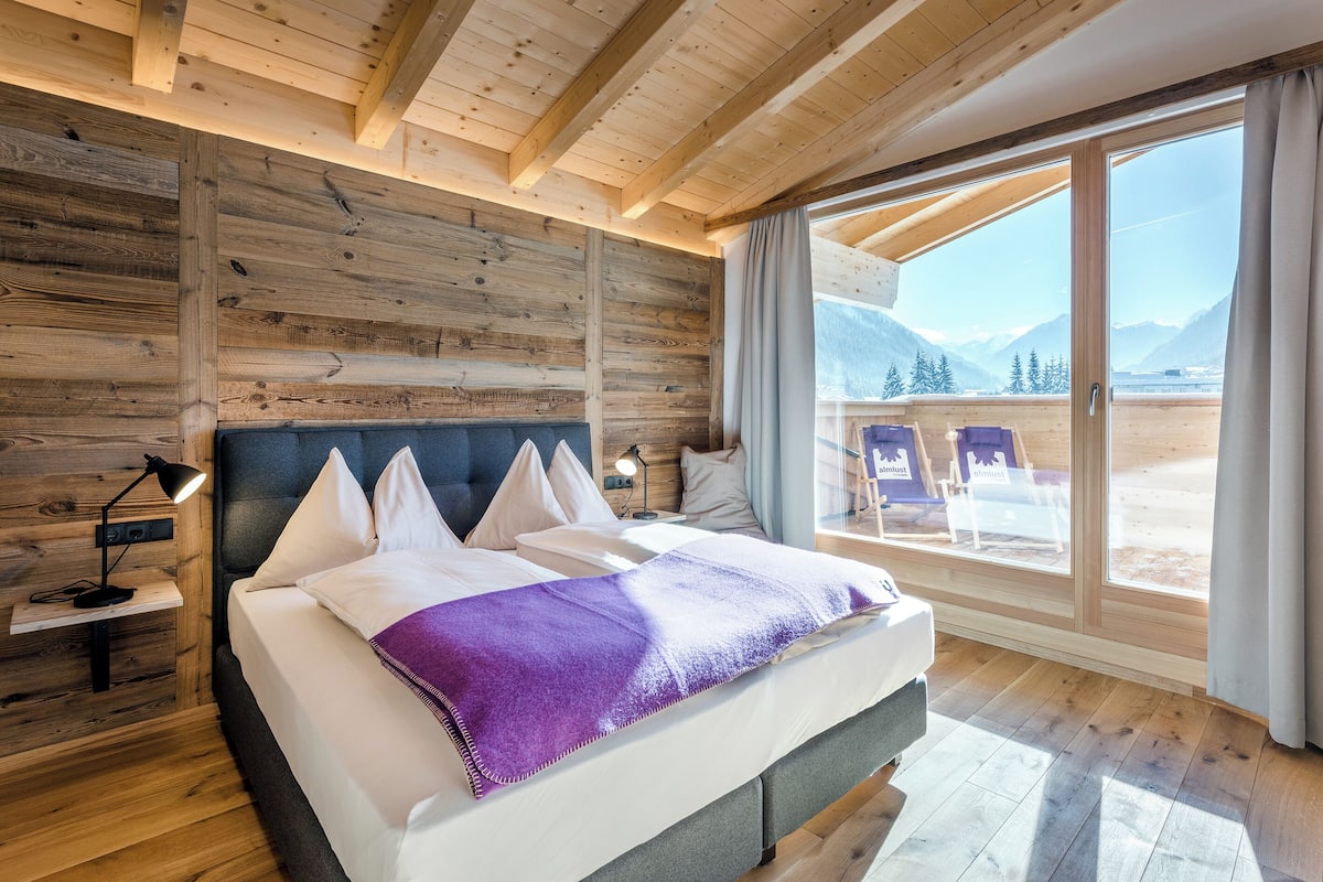 A bedroom with a bed, wooden walls, a nightstand, a lamp, and a large window with a mountain view.