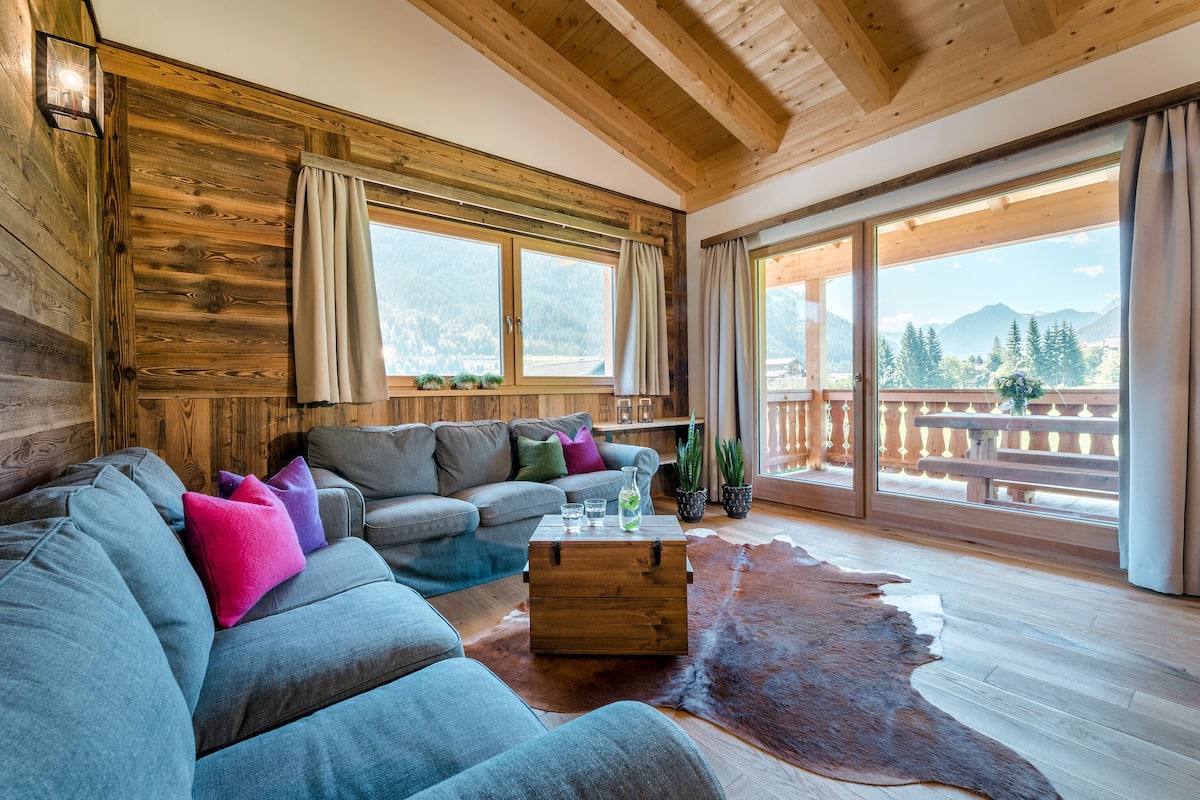 A cozy living room with a wooden ceiling, large windows, a leather sofa, a wooden coffee table, and a cowhide rug.