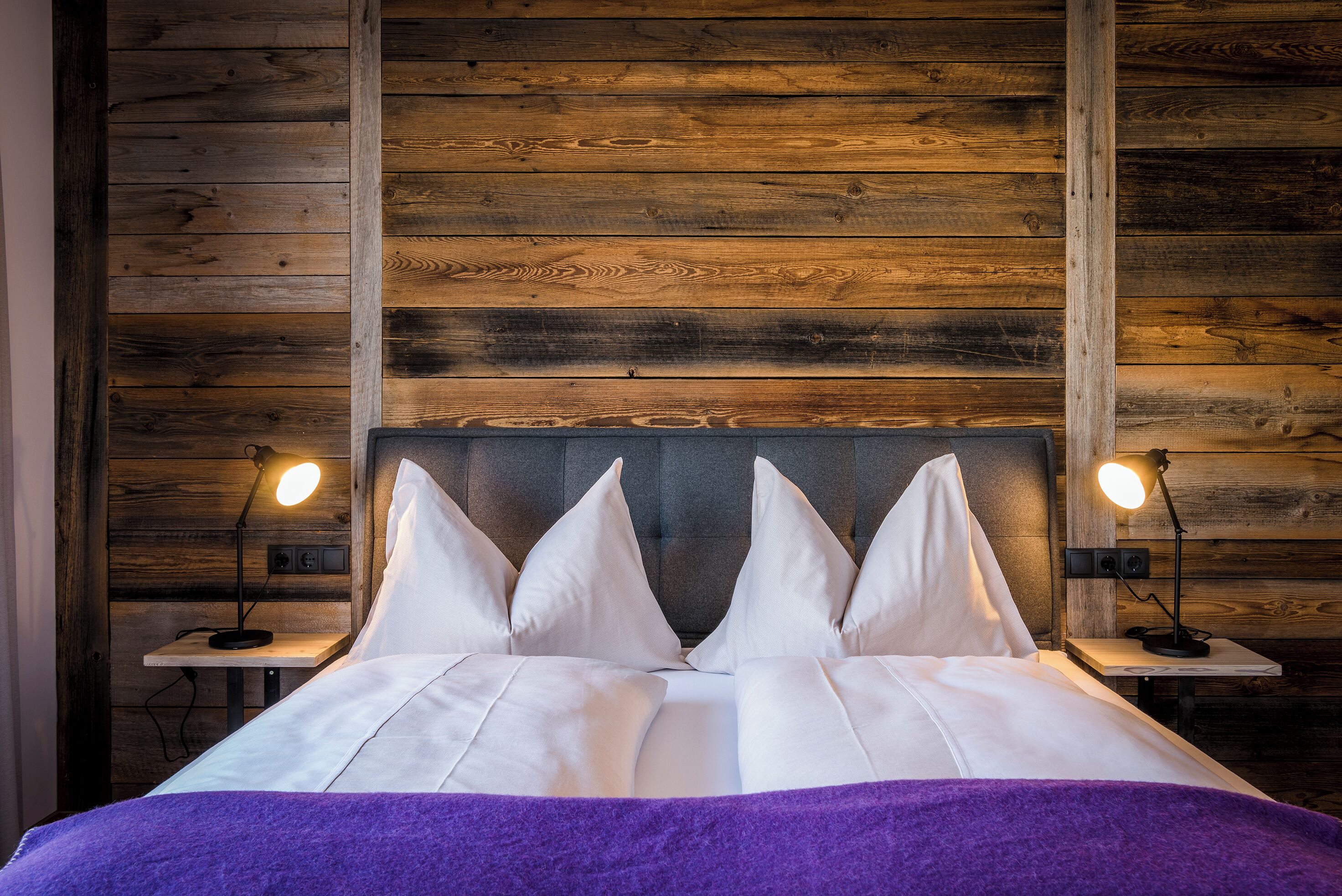 A bed with white linens and a purple throw, flanked by two bedside tables with lamps, set against a wooden wall.