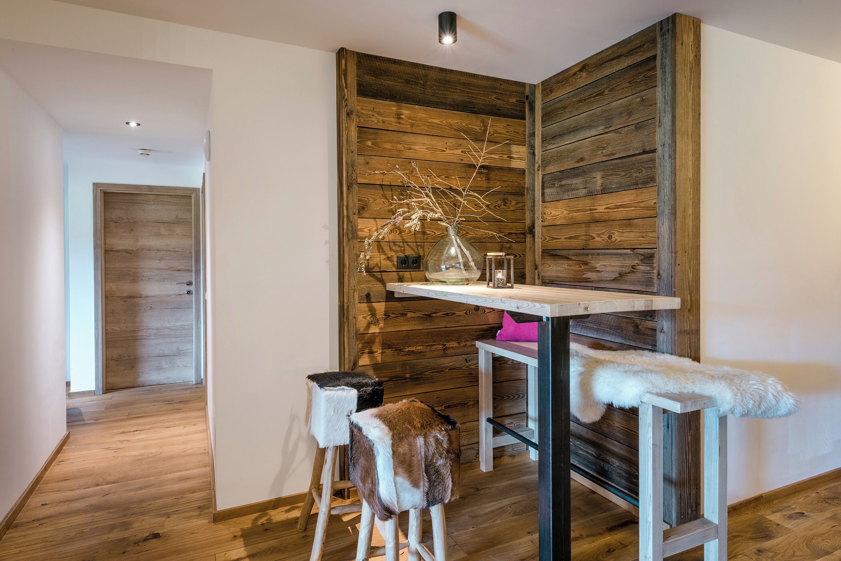 Una habitación con una pared de acento de madera, una pequeña mesa con un florero, y dos taburetes con mantas de piel.