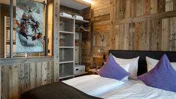 A bedroom with a wooden wall, a bed with white bedding and purple pillows, a wardrobe, and a painting of a group of people on a raft.
