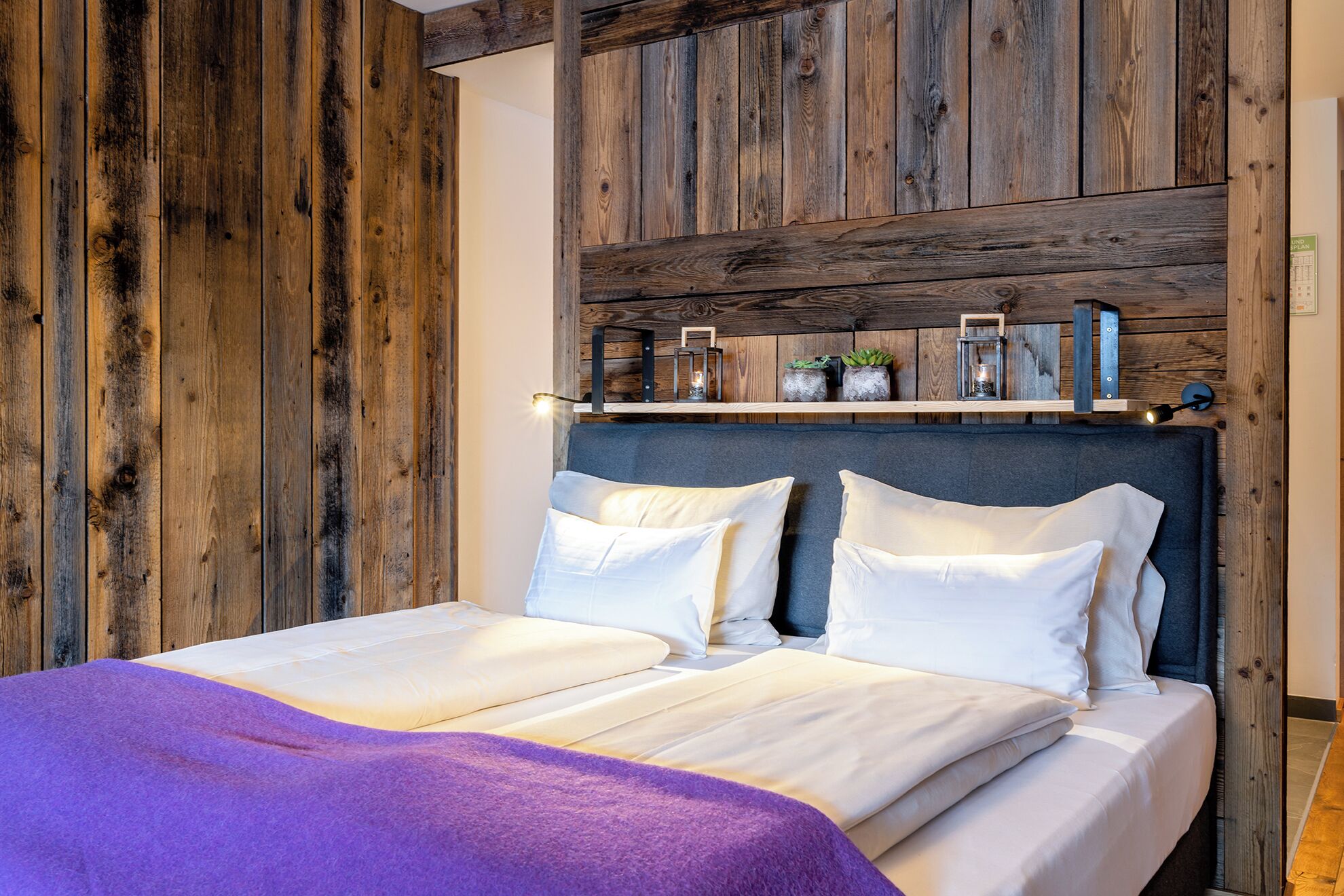 A double bed with white linens, a purple throw blanket, and a wooden headboard with shelves above.