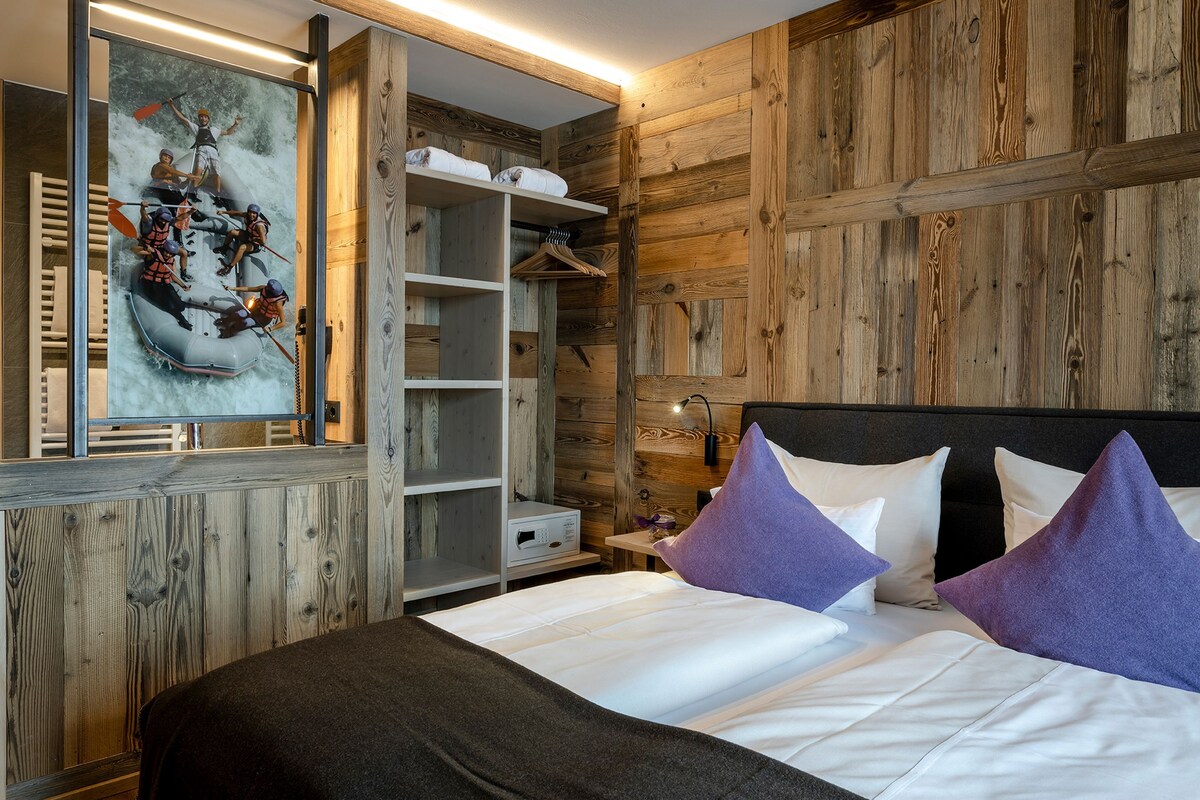 Quarto com parede de madeira, cama com roupa de cama branca e almofadas roxas, guarda-roupa e pintura de um grupo de pessoas em uma jangada.