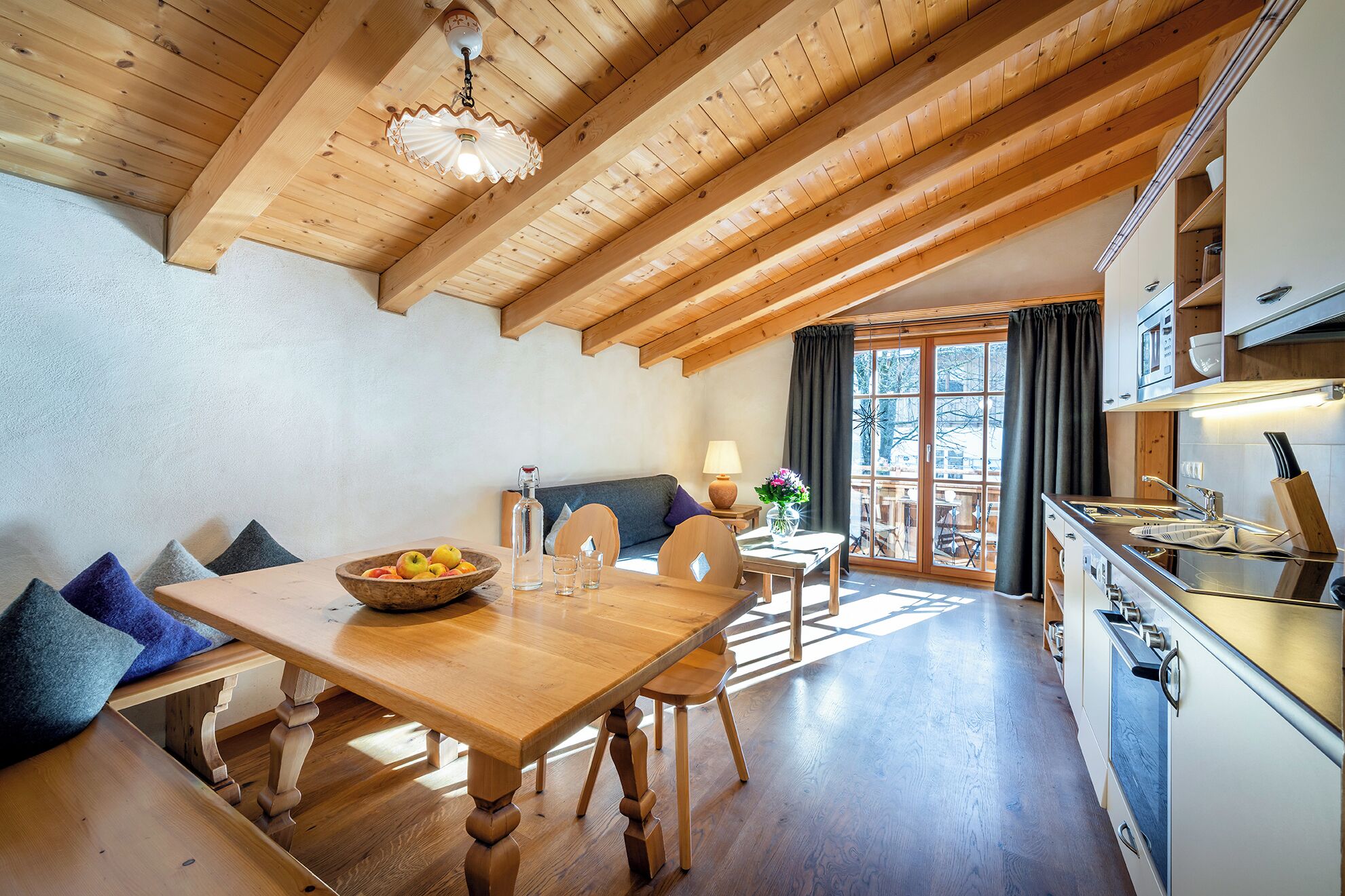 A wooden dining table with chairs, a bowl of fruit, a lamp, and a view of the outdoors through a window.