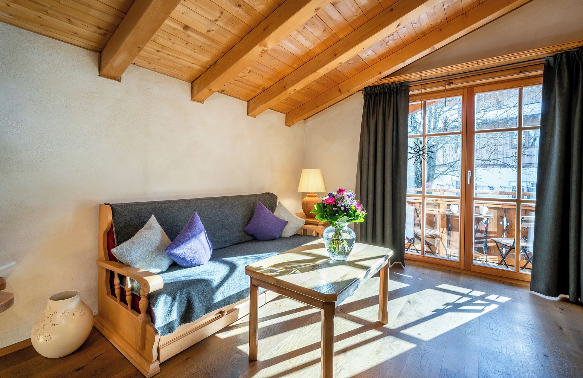 Una habitación acogedora con techo de madera, un sofá con cojines, una mesita con un florero y un ventanal con cortinas.