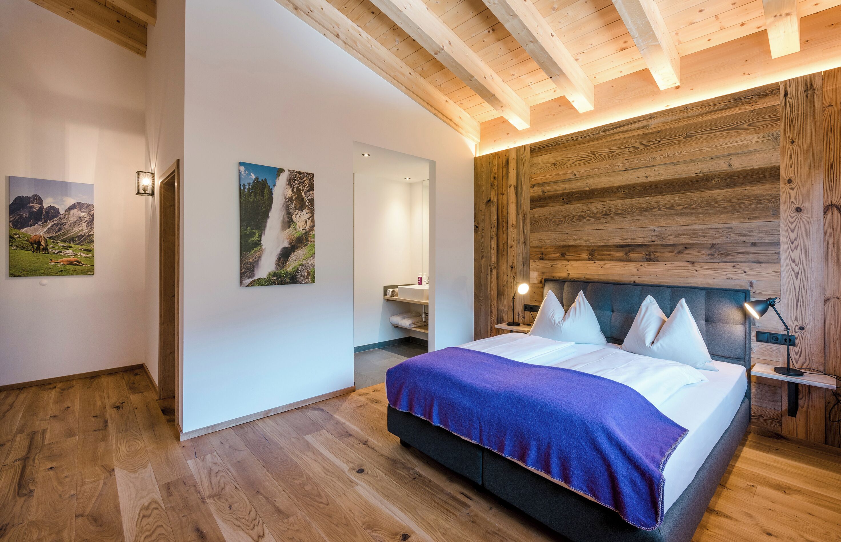 A bedroom with a wooden ceiling, a bed with white and purple bedding, and two wall-mounted light fixtures.