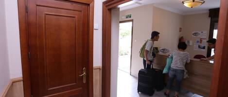 A hotel reception area with a wooden door labeled "CONSIGNA," a reception desk, and a sign indicating the "SALIDA EXIT."