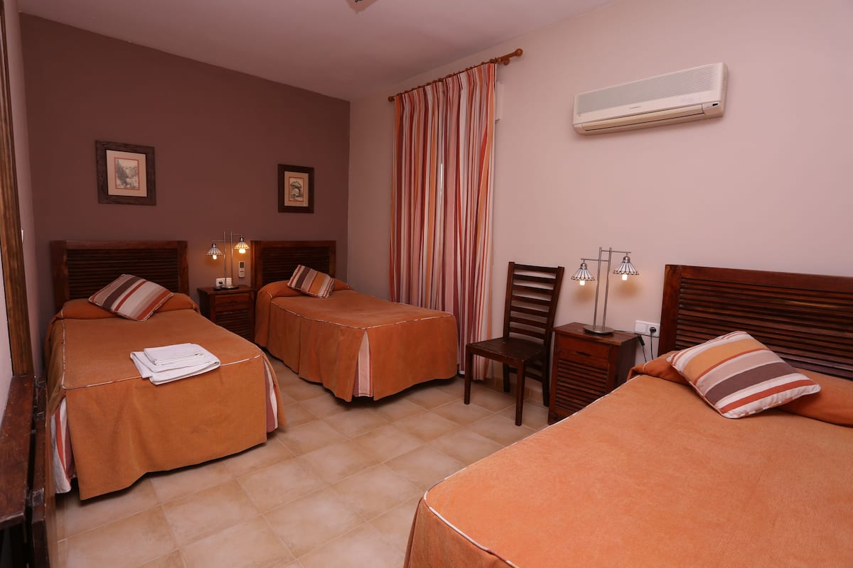 A hotel room with three beds, a wooden headboard, a chair, a nightstand, and an air conditioning unit.