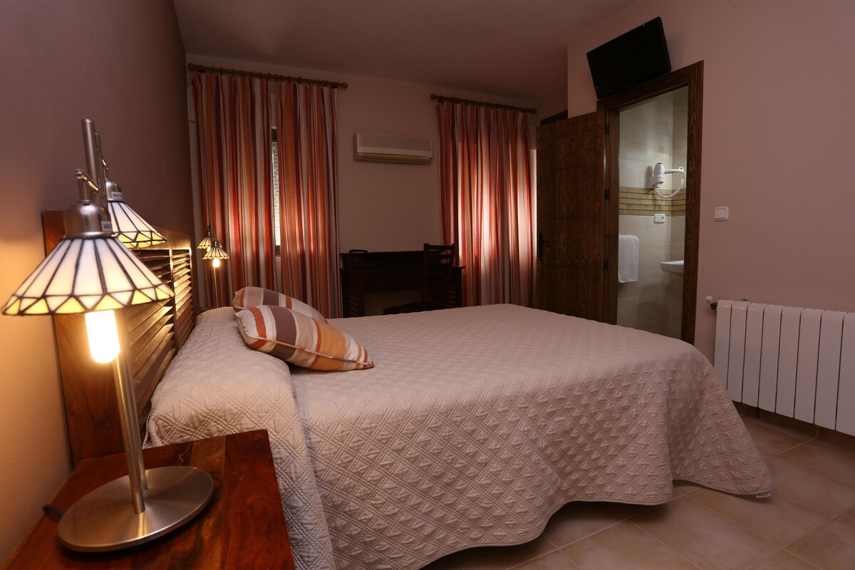 Un dormitorio con cama, mesita de noche con lámpara, una silla y un baño visible a través de una puerta abierta.