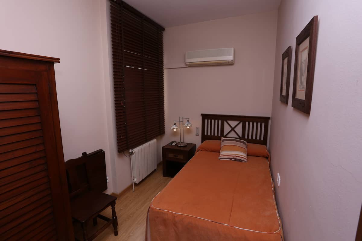 A small, narrow room with a single bed, a wooden chair, a bedside table with a lamp, and a radiator.
