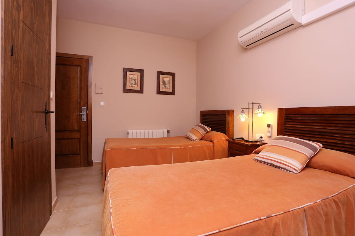 A hotel room with two beds, a wooden wardrobe, an air conditioning unit, and two framed pictures on the wall.