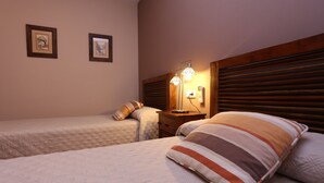 Habitación de hotel con dos camas, cabecera de madera, mesita de noche con lámpara y cuadros en la pared.