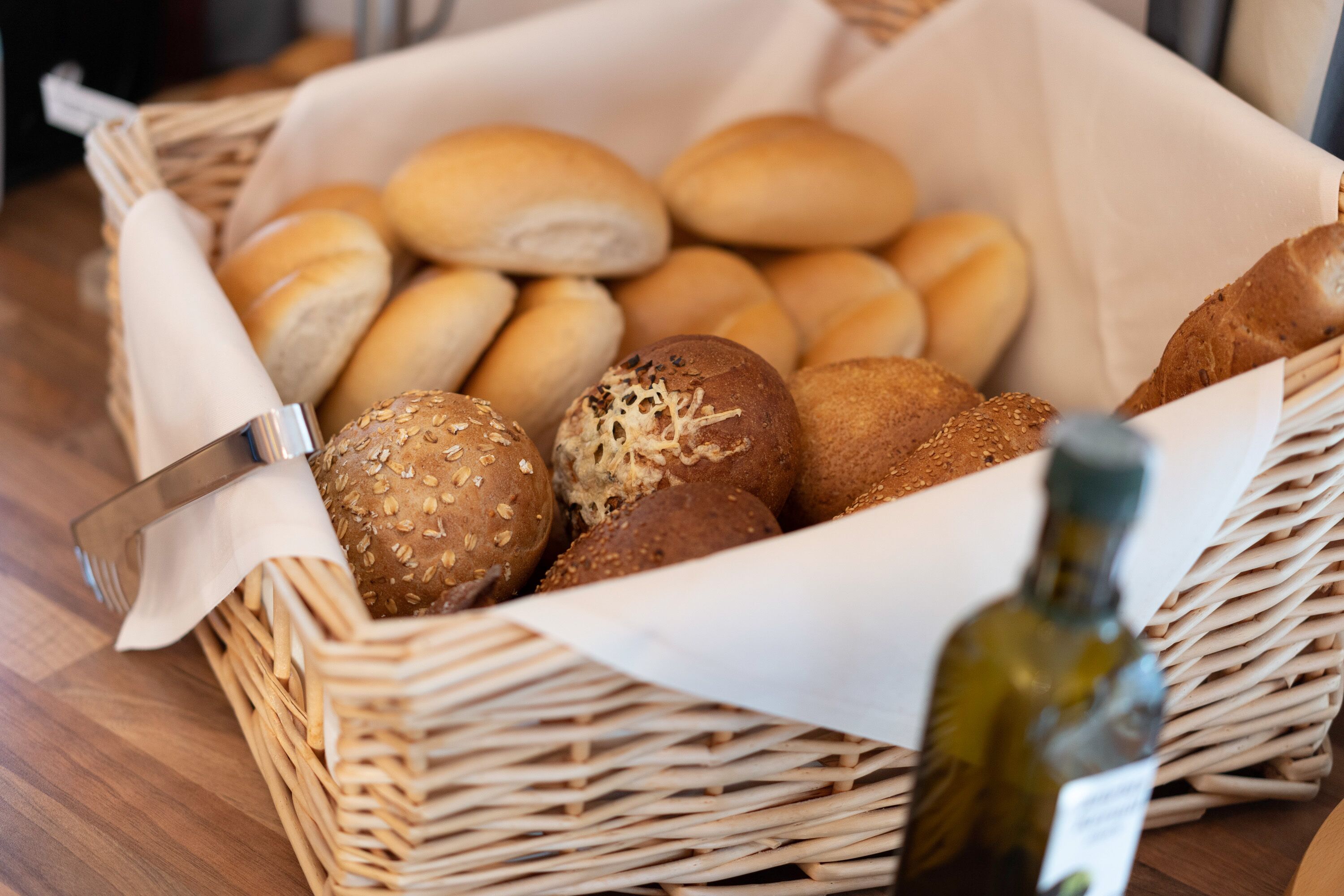 A wicker basket filled with various types of bread, including rolls and seeded buns, placed on a wooden table.