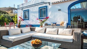 Un edificio blanco con detalles azules, un sofá de mimbre con cojines, una mesa de vidrio con frutero, y vistas del paisaje.