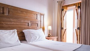 Habitación de hotel con cabecera de madera, cama con ropa blanca, mesita de noche con lámpara y vista a través de una puerta abierta.