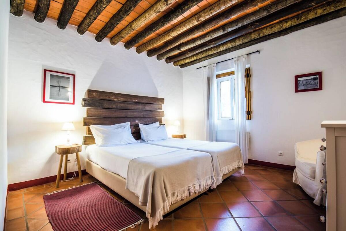 Une chambre avec un plafond en bois, un lit recouvert de linge de lit blanc, un tapis rouge et une petite table de chevet avec une lampe.