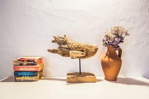 Una escultura de pescado de madera sobre un soporte, una pila de libros y un jarrón con flores secas sobre una superficie blanca.