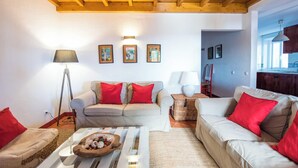 Una sala de estar acogedora con techo de madera, dos sofás con cojines rojos, una mesa de centro con frutero y una lámpara.