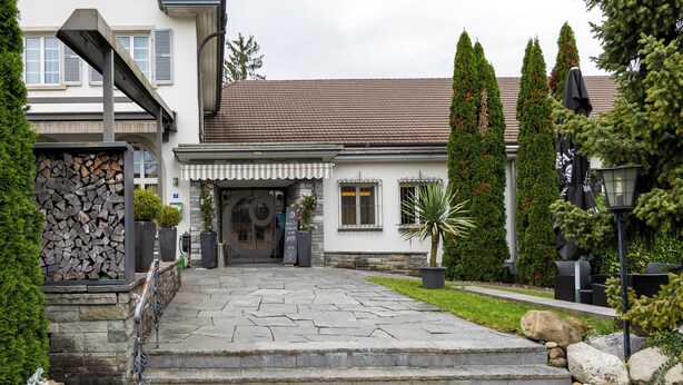 A stone staircase leading to a building with a tiled roof, a stone wall with logs, and a well-maintained lawn with rocks and plants. - Hotel & Gasthaus die Perle (Perlen)