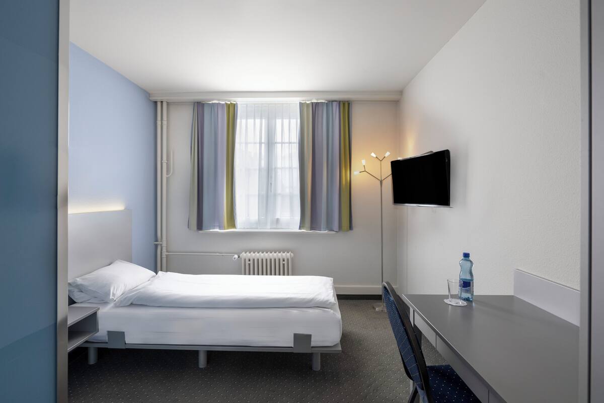 A hotel room with a bed, a desk, a chair, a TV, and a window with curtains.