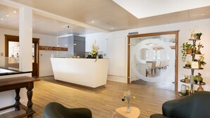 A reception area with a wooden reception desk, a green sofa, a small round table with a vase, and a plant shelf. - Hotel & Gasthaus die Perle (Perlen)