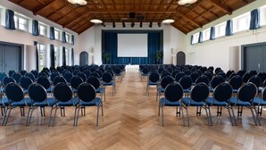 A conference room with rows of chairs, a stage with a screen, wooden ceiling beams, and large windows. - Hotel & Gasthaus die Perle (Perlen)