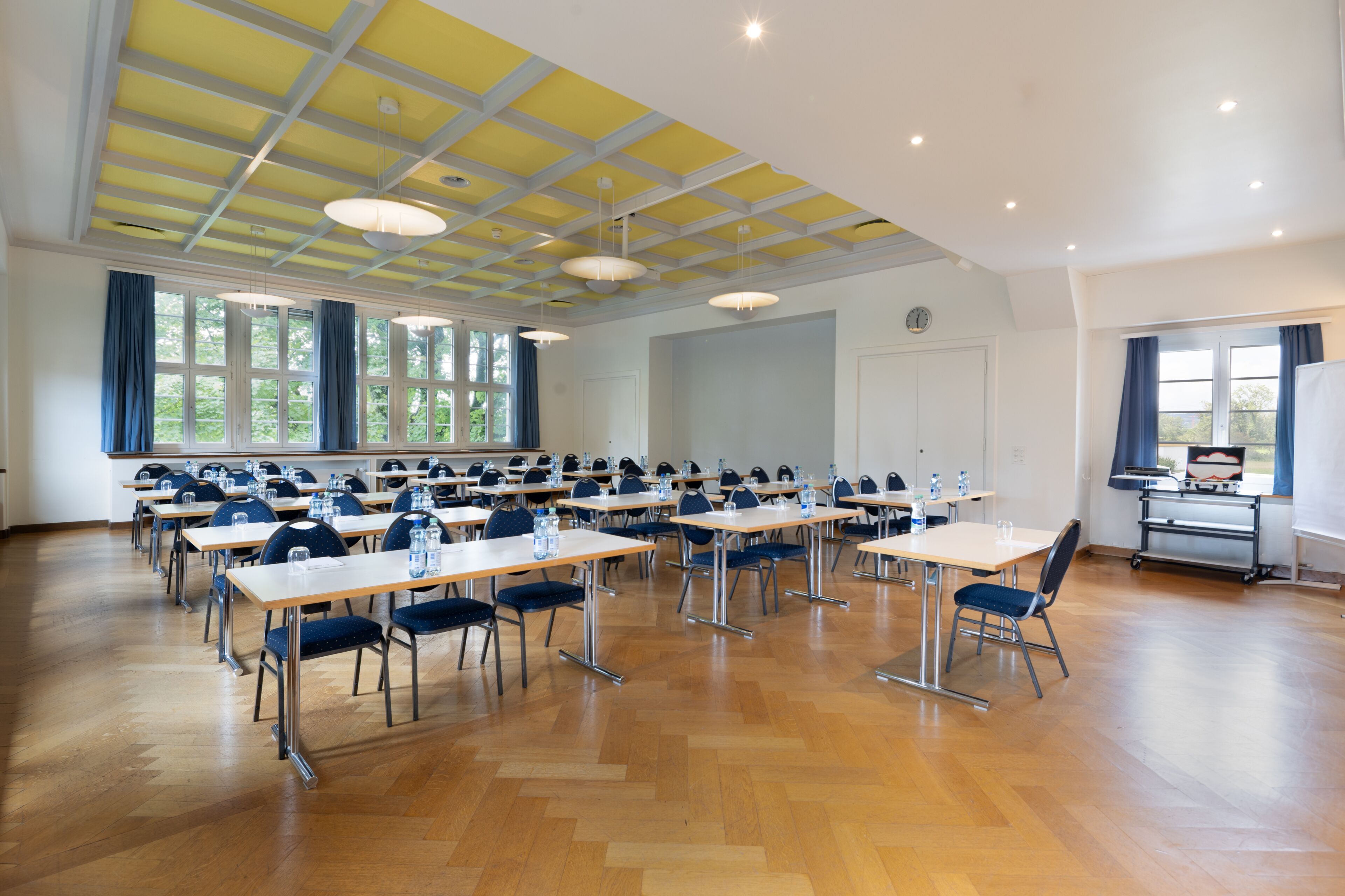 A large conference room with multiple tables and chairs, large windows, and a high ceiling with exposed beams.