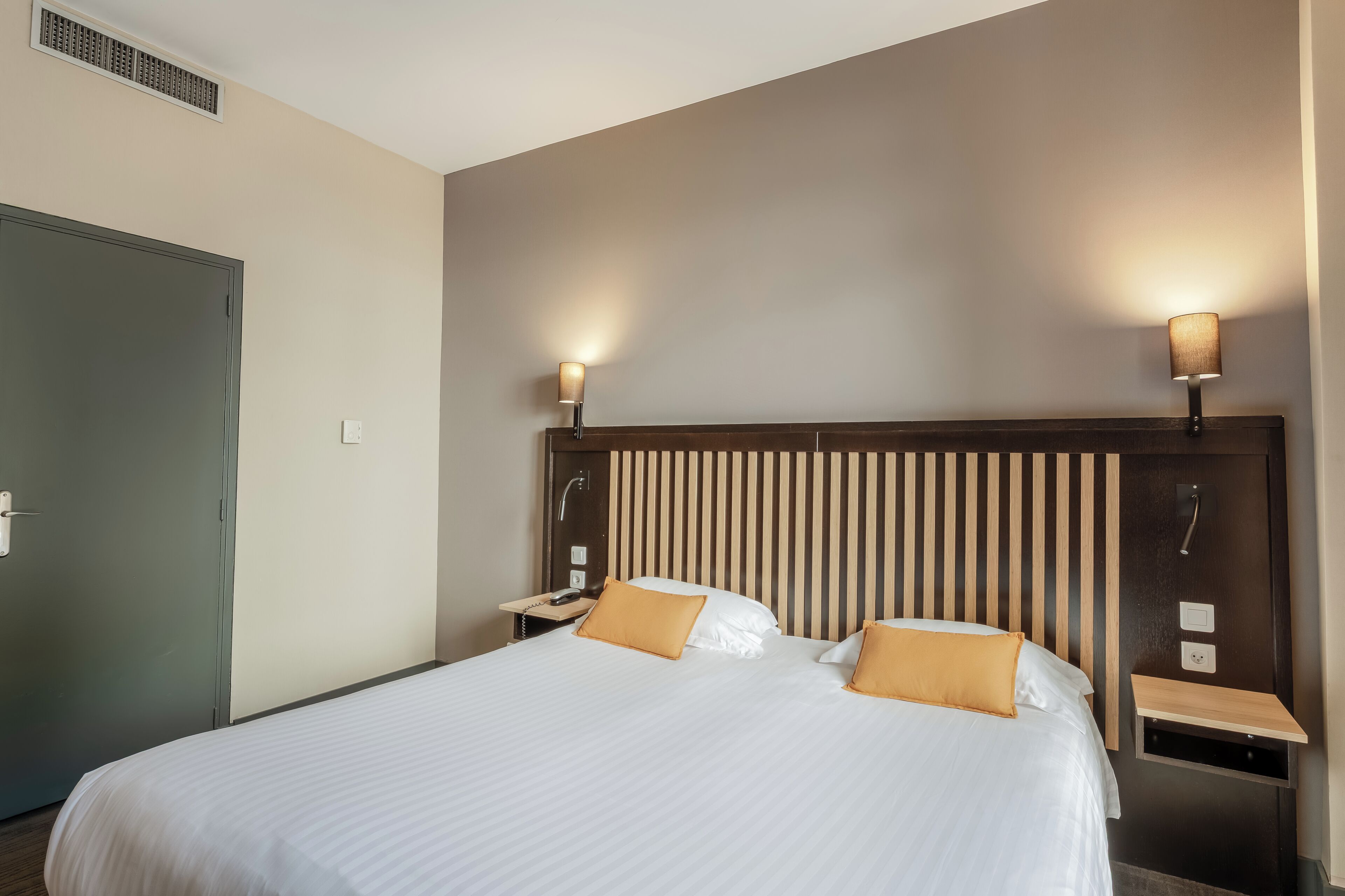 Habitación de hotel con una cama grande, dos mesitas de noche, un cabecero de madera y dos lámparas de pared.