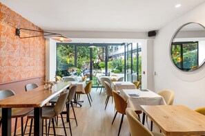 An indoor dining area with tables and chairs, a large window with a view of the outdoors, and a wall with a leaf pattern. - Hotel Molière (Cannes)