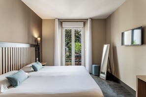 A hotel room with a bed, a bedside lamp, a TV, a window with a view of greenery, and a door leading to an outdoor area. - Hotel Molière (Cannes)