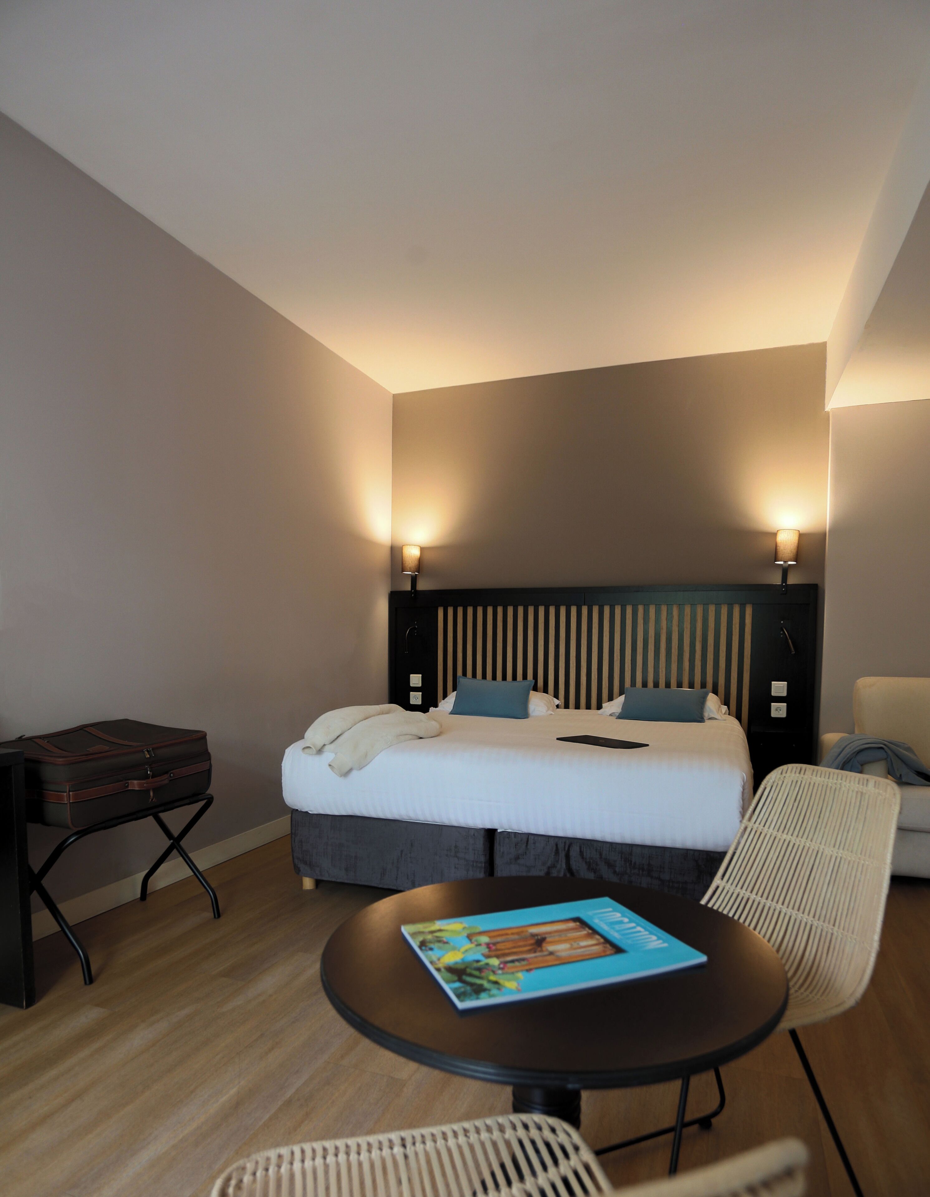 Habitación de hotel con cama, lámparas de noche, una mesa con un libro, una silla y una maleta.