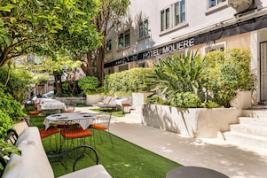 Un área de estar al aire libre con mesas y sillas, rodeada de follaje y un edificio con un letrero que dice 'HOTEL MOLIERE'.
