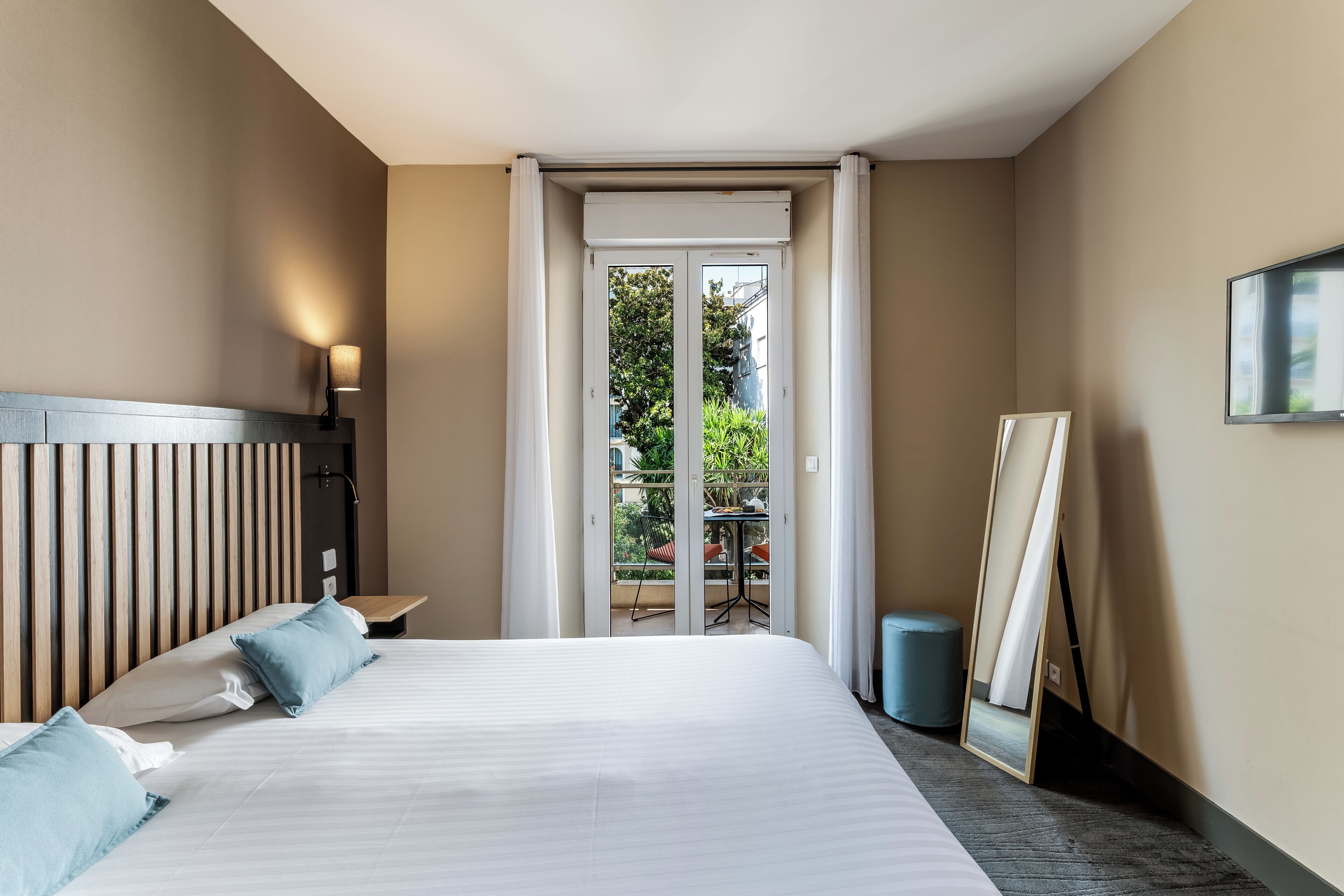 Ein Hotelzimmer mit Bett, Nachttisch, Lampe, Spiegel und Blick auf den Balkon.